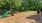 Small image of slide and playground objects on a floor of wood chips. Green bushes behind the park. 